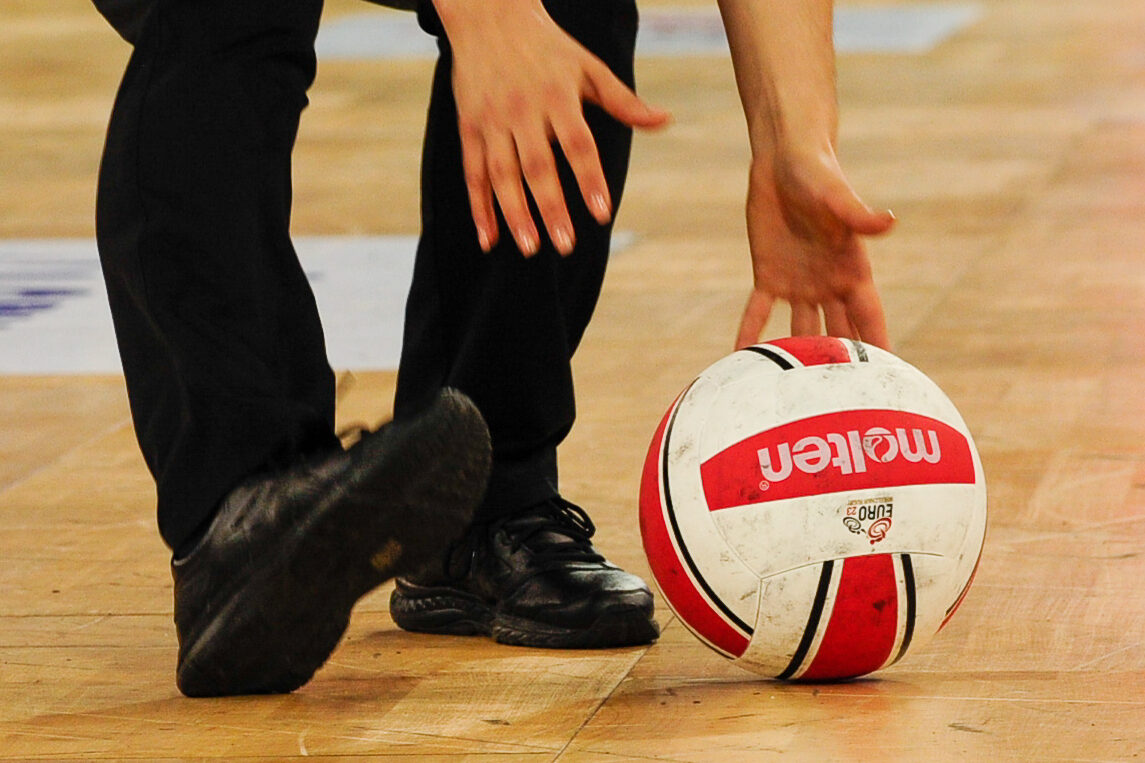 Ein Schiedsrichter hebt den Spielball vom Boden auf. Zu sehen sind zwei Hände, der Ball und daneben die Beine des Referees.